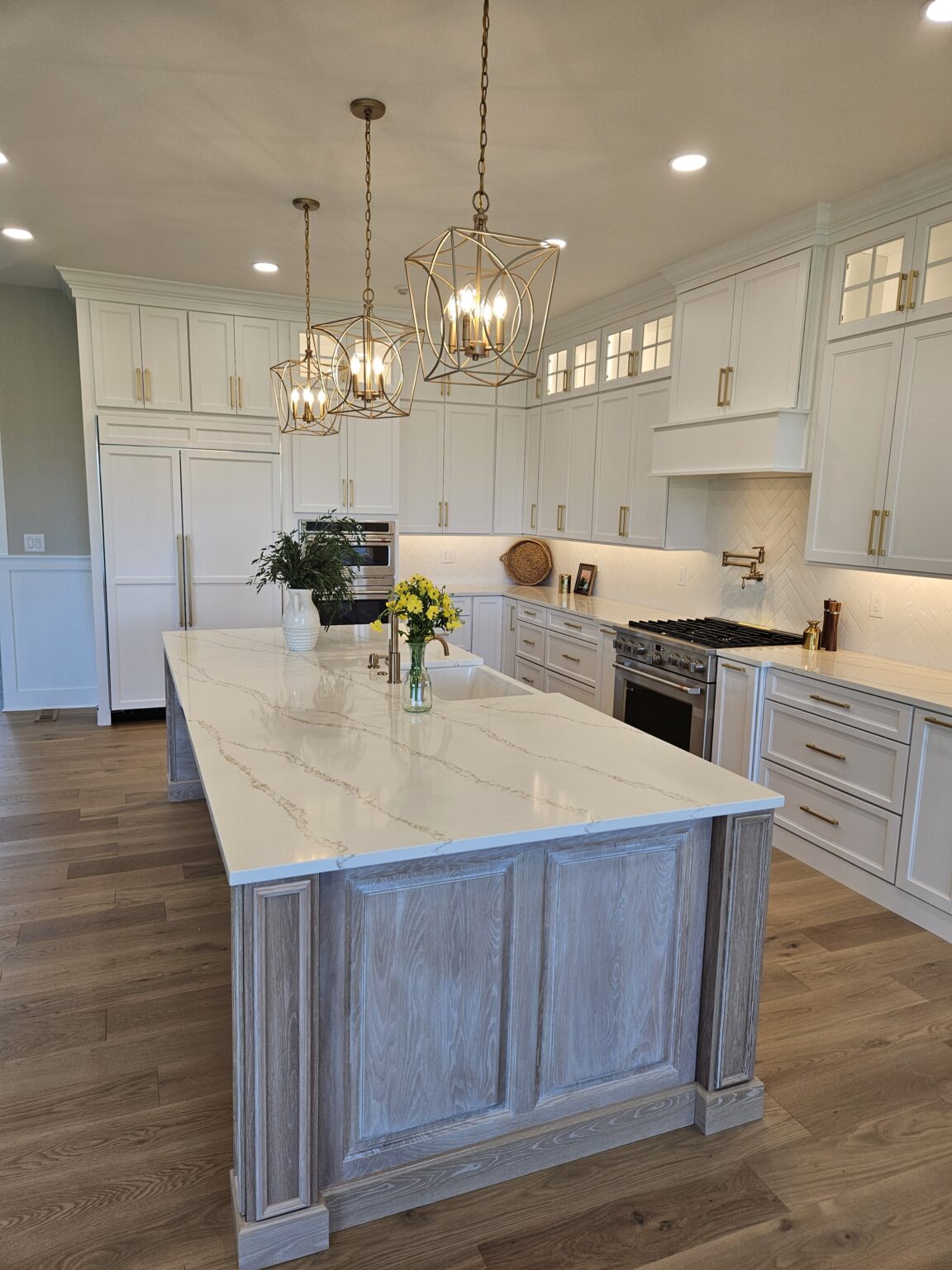 Integrity Cabinets custom kitchen featuring a cerused driftwood island with quartz countertops, white shaker perimeter cabinets, and gold cage pendant lights.