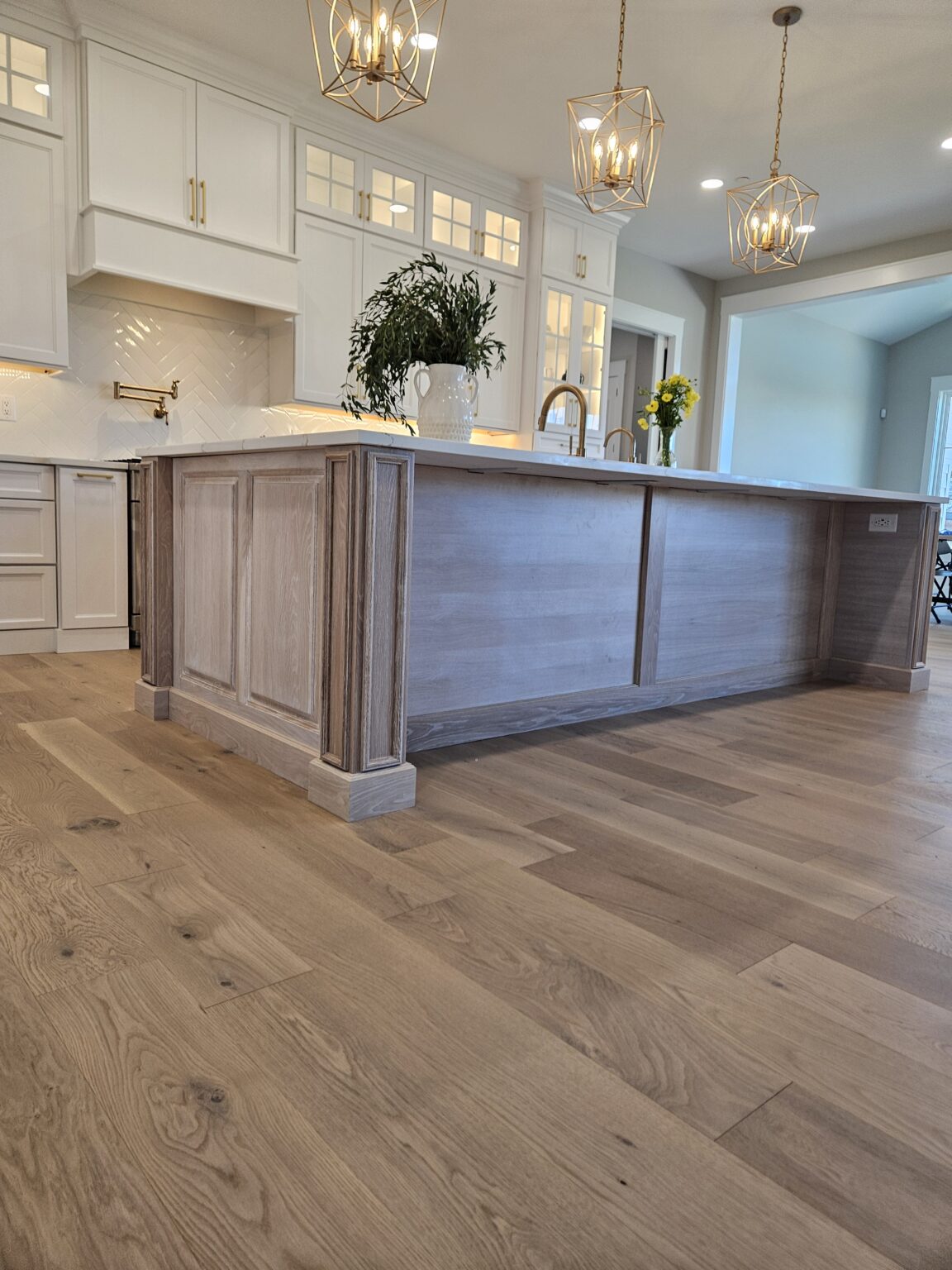 Integrity Cabinets custom kitchen island in cerused driftwood finish with decorative corner posts, white perimeter cabinets, and gold pendant lights.