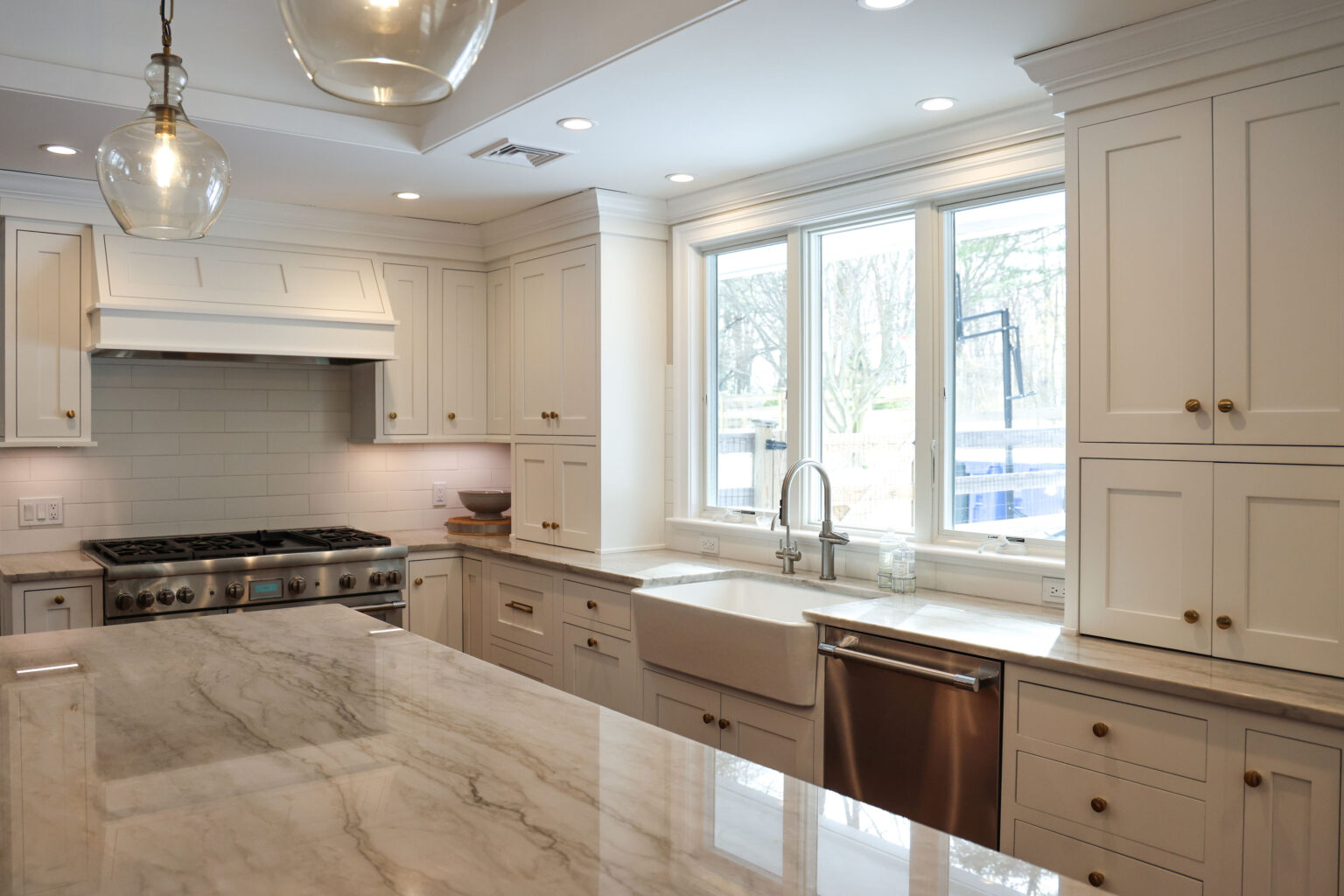 Custom white Shaker-style Brighton cabinetry in a luxury kitchen with brass hardware, polished marble countertops, a farmhouse sink under wide windows, and hanging glass pendant lights.