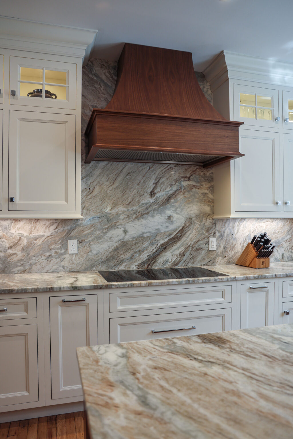 Luxury custom kitchen featuring cream Brighton Cabinetry with glass-front uppers, a striking wood range hood, and exotic granite countertops with a matching full-height backsplash.