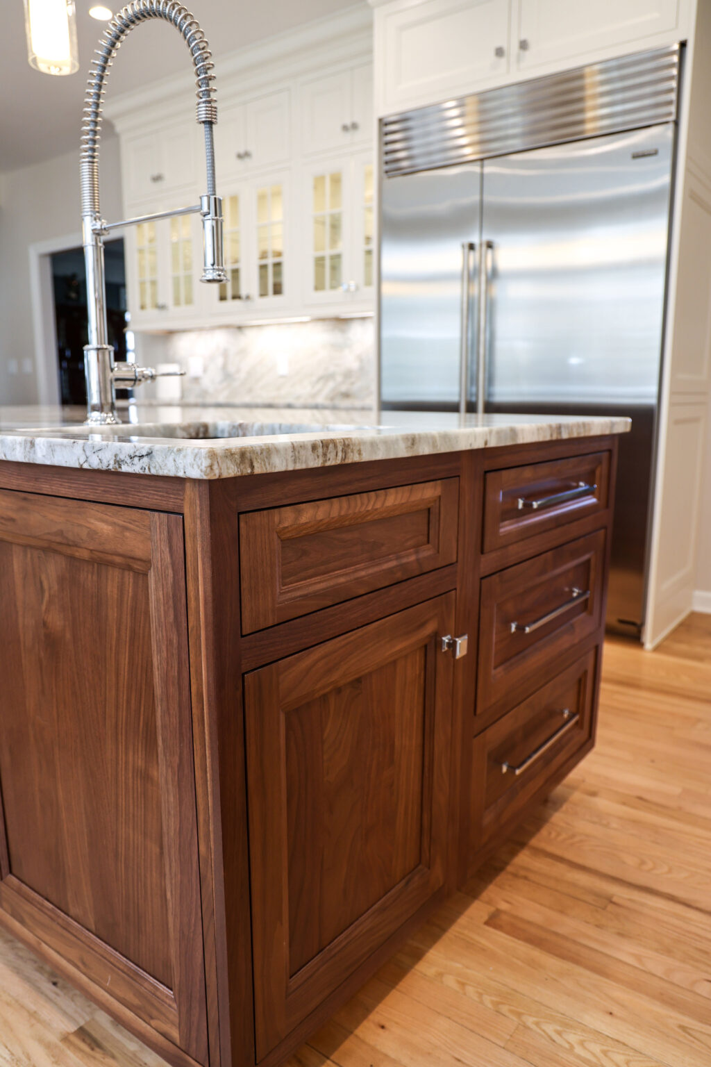 Custom kitchen island with walnut wood Brighton cabinetry, natural stone countertops, and a modern coiled spring faucet, contrasting with white upper cabinets and a professional built-in refrigerator.